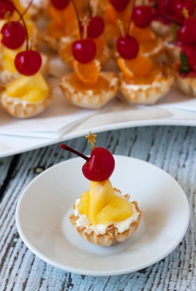Creamy no-bake coconut cheesecake in a crisp, flaky mini fillo shell topped with a sweet pineapple topping and garnished with fresh pineapple and a maraschino cherry. 