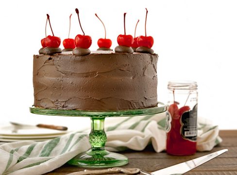 Side view of a fully baked and decorated old-fashioned chocolate cake with cherry filling, glossy chocolate icing and maraschino cherries on top, next to a jar of cherries and a green and white striped cloth.