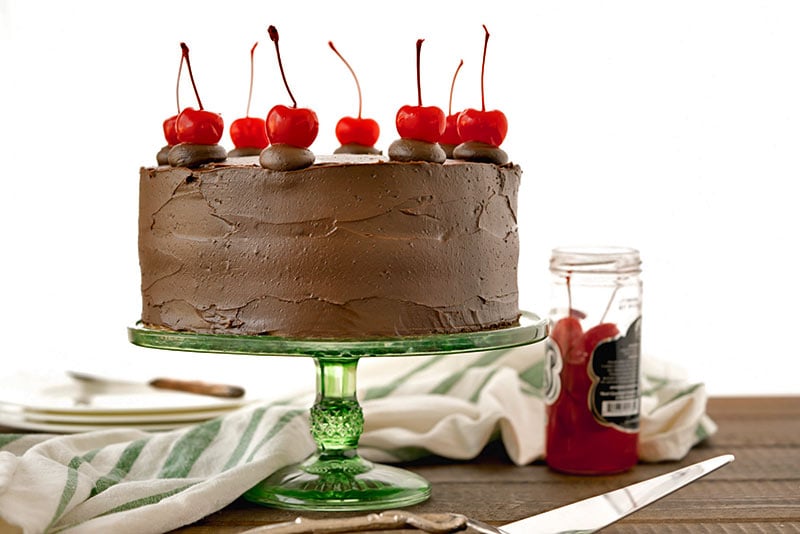 Side view of a fully baked and decorated old-fashioned chocolate cake with cherry filling, glossy chocolate icing and maraschino cherries on top, next to a jar of cherries and a green and white striped cloth.