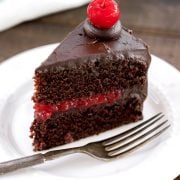 A single slice of old-fashioned chocolate cake filled with homemade cherry filling and frosted with easy chocolate icing, topped with a stemmed maraschino cherry on a white dessert plate with a silver fork.