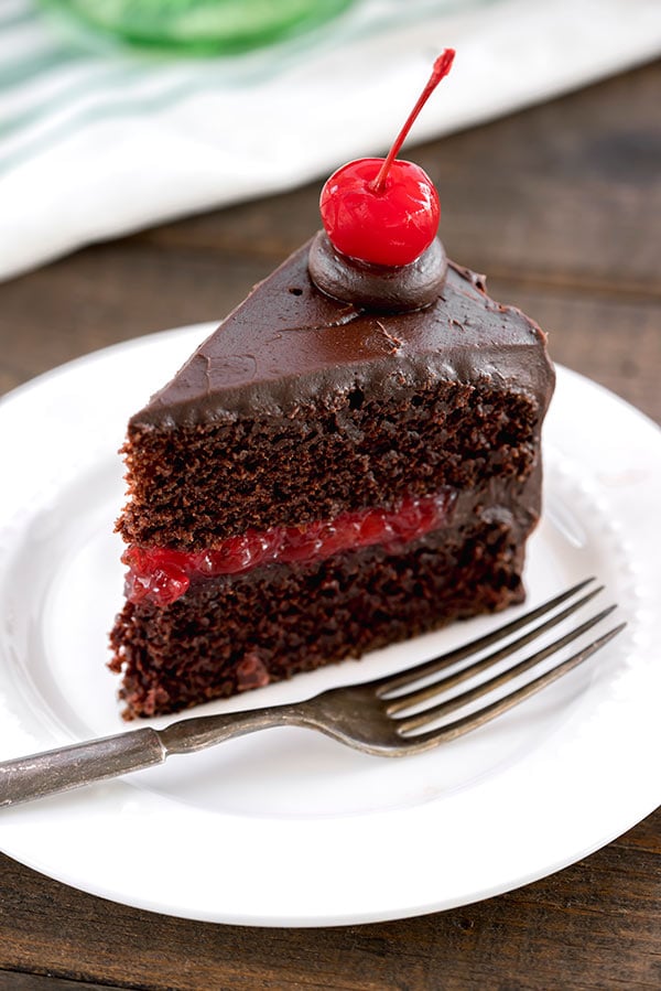 A single slice of old-fashioned chocolate cake filled with homemade cherry filling and frosted with easy chocolate icing, topped with a stemmed maraschino cherry on a white dessert plate with a silver fork.