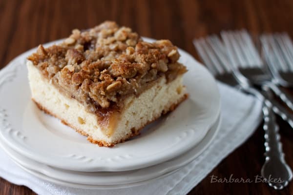 Holiday Apple Kuchen A square slice of crumb-topped apple kuchen coffee cake sits on a white plate with a scalloped edge. The plate rests on a white napkin, with several forks in the background on a wooden table.