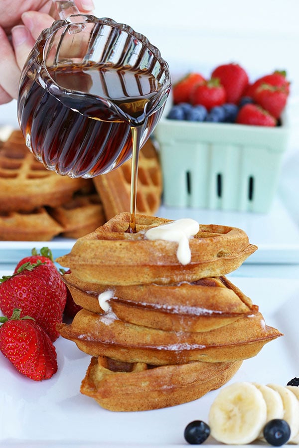 Syrup being poured onto a stack of whole wheat waffles in quarters on a plate ready to eat.