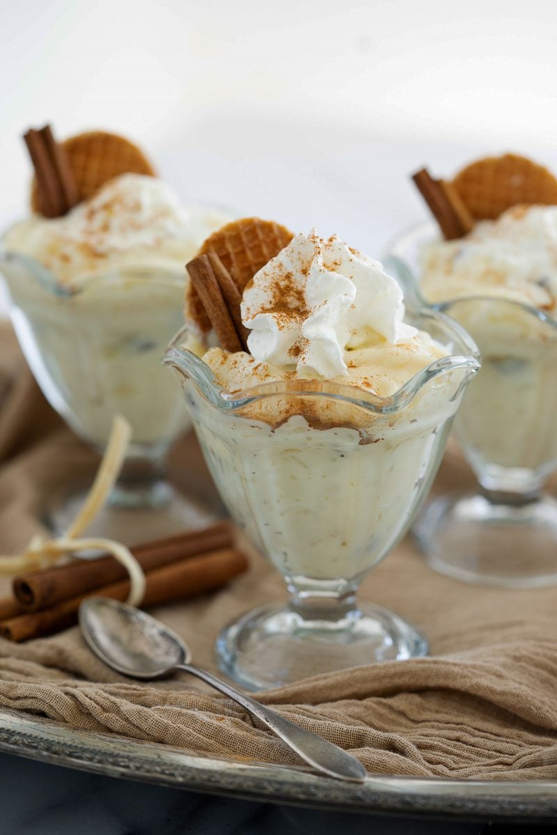 Close up on three glass goblets of homemade old-fashioned white rice pudding flavored with cinnamon and topped with whipped cream and waffle cookies.