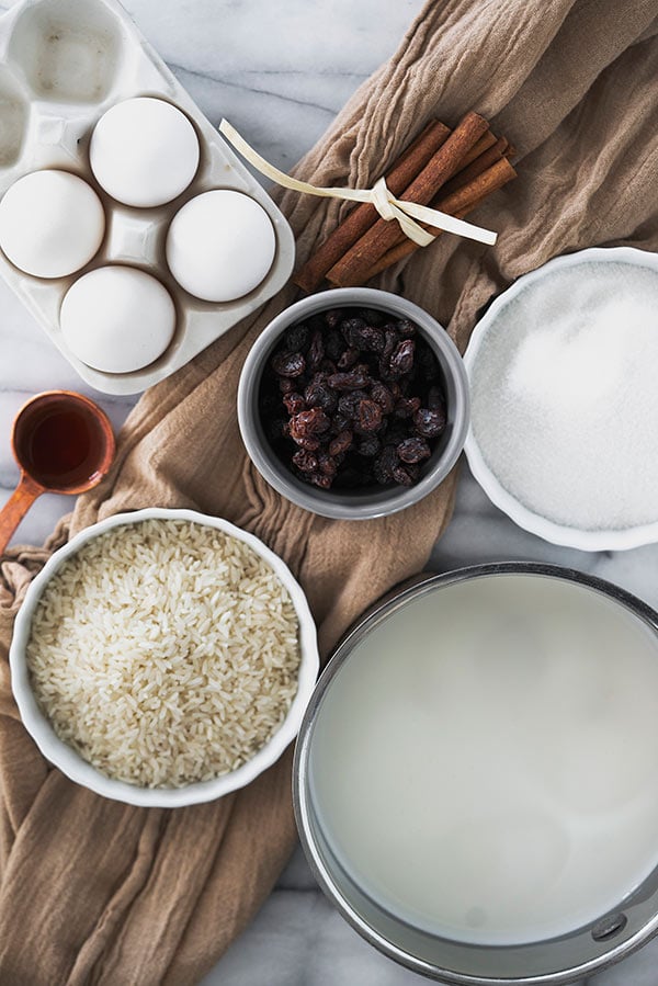 Overhead of ingredients for Old-fashioned rice pudding: eggs, raisins, long-grain white rice, vanilla extract, milk, sugar and cinnamon on a white marble background with a brown linen.