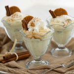 Side view of three ice cream cups filled with homemade old-fashioned rice pudding topped with cinnamon, whipped cream and waffle cookies on a brown linen cloth with cinnamon sticks and more cookies.