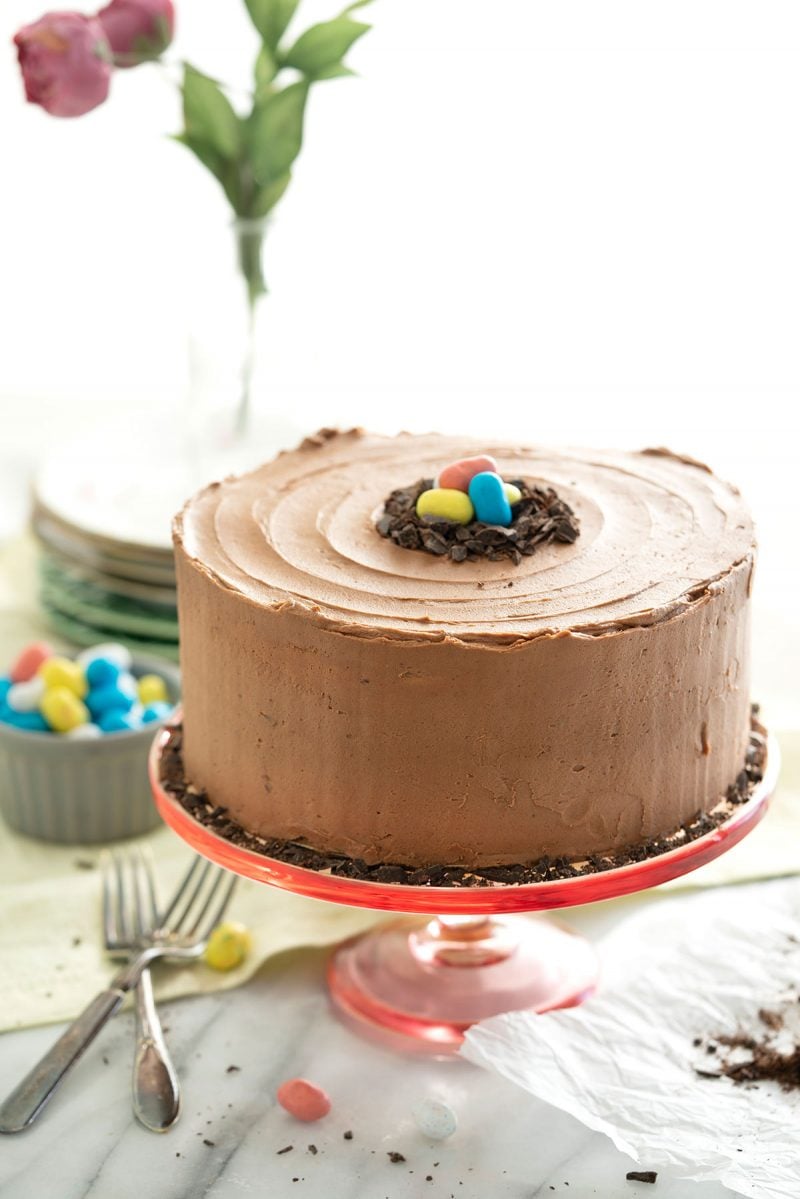 Malted milk chocolate layer cake on a red cake stand frosted in smooth fluffy homemade chocolate frosting and decorated with colorful candy.
