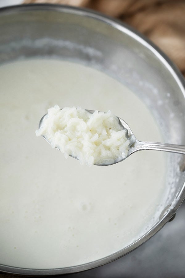 Close up of a teaspoon of perfectly cooked long-grain white rice over a saucepan of milk and rice cooking for old-fashioned rice pudding recipe.