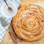 Overhead of beautiful rose-shaped cinnamon roll coffee cake with cinnamon sugar coating and sugar glaze on a light wooden background with a blue and white checkered cloth.