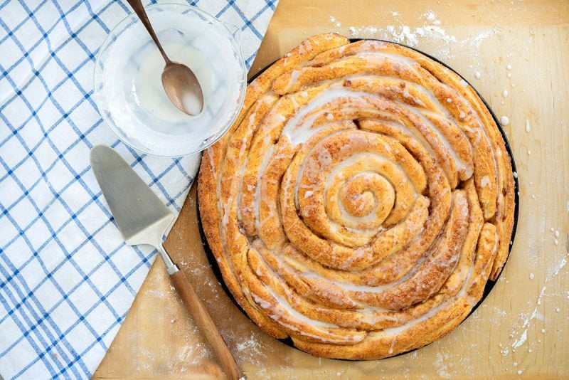 Overhead of beautiful rose-shaped cinnamon roll coffee cake with cinnamon sugar coating and sugar glaze on a light wooden background with a blue and white checkered cloth.