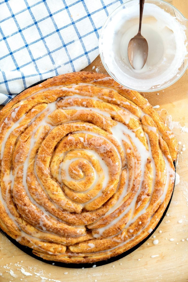 Overhead of a homemade cinnamon roll coffee cake with a white sugar glaze on a wooden background with a white and blue checkered cloth.