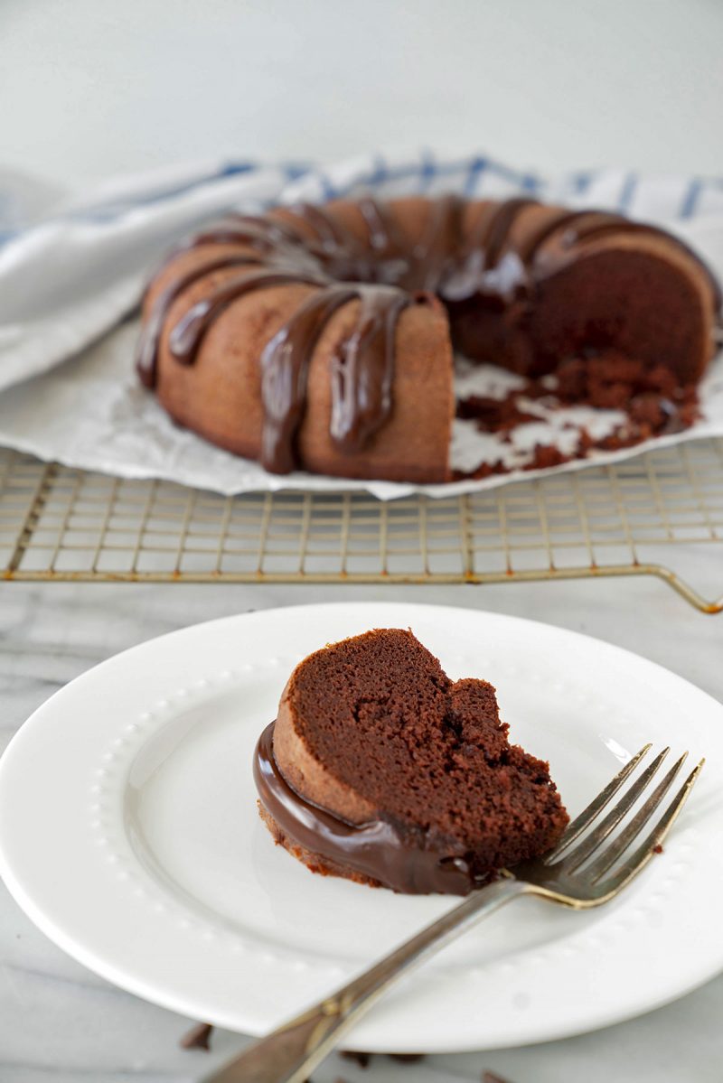 A slice of chocolate bundt cake with drizzle of chocolate frosting on a plate with cake in the background
