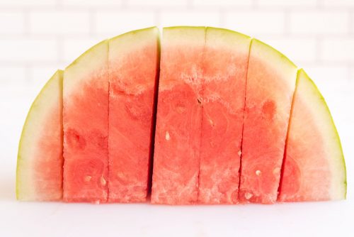 one half of a red watermelon on a cutting board flseh side down sliced into equal triangles before cutting into cubes