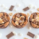 Three S'mores Brookie Bowls in a row surrounded by mini marshmallows, Hershey's bars and broken graham crackers