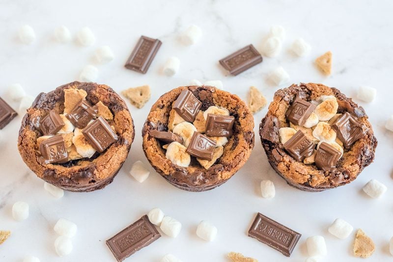 Three S'mores Brookie Bowls in a row surrounded by mini marshmallows, Hershey's bars and broken graham crackers