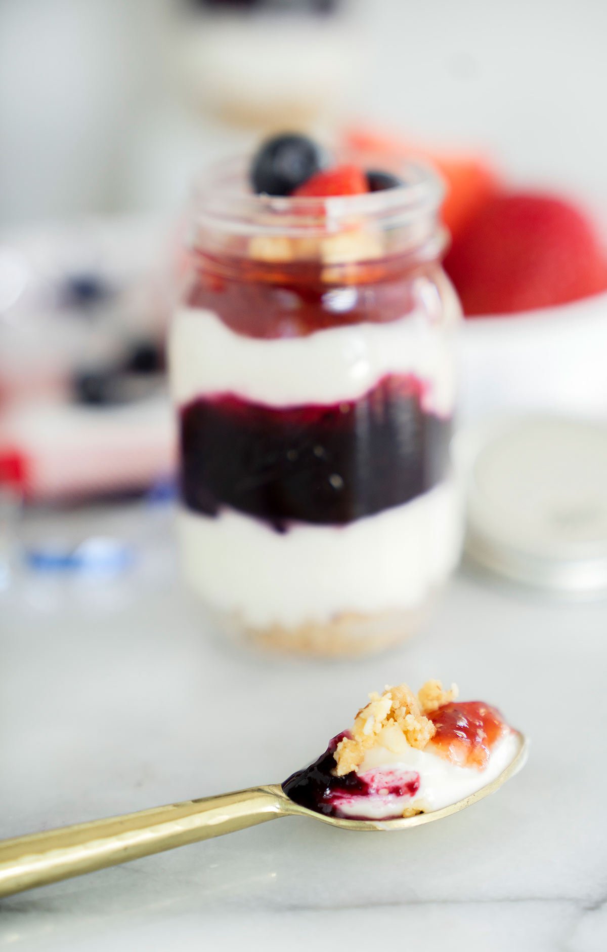 red white and blue cheesecake in a mason jar with fresh blueberry and strawberry compote, cream cheese filling and cookie crust with a spoonful in front