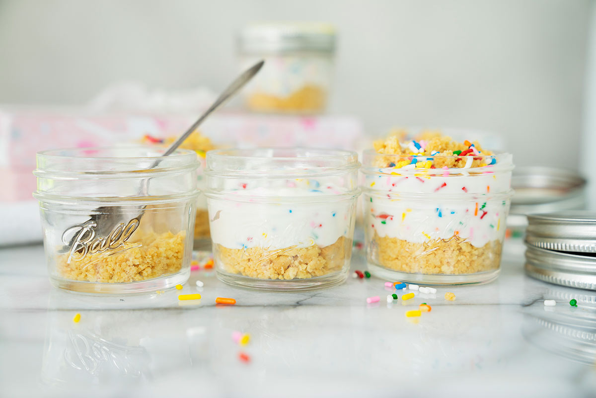 three four ounce mason jars being filled with no-bake birthday cake cheesecake