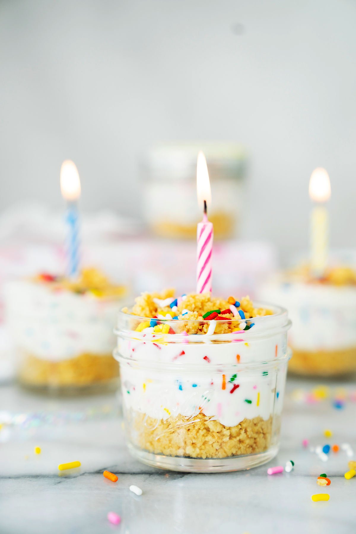 birthday cake cheesecake in a jar with no-bake oreo cookie crust and creamcheese filling with rainbow sprinkles and a birthday candle