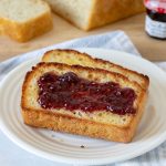 Two slices of English muffin bread, one with jam, on a white plate. A loaf of English muffin bread on a cutting board with raspberry jam in the background.