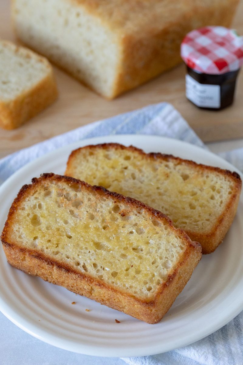 Two slices of buttered English muffin bread on a white plate. A loaf of English muffin bread on a cutting board with raspberry jam in the background.