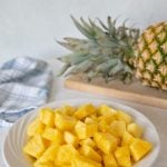 cut up pineapple chunks in a white bowl with a whole pineapple on a cutting board in the background