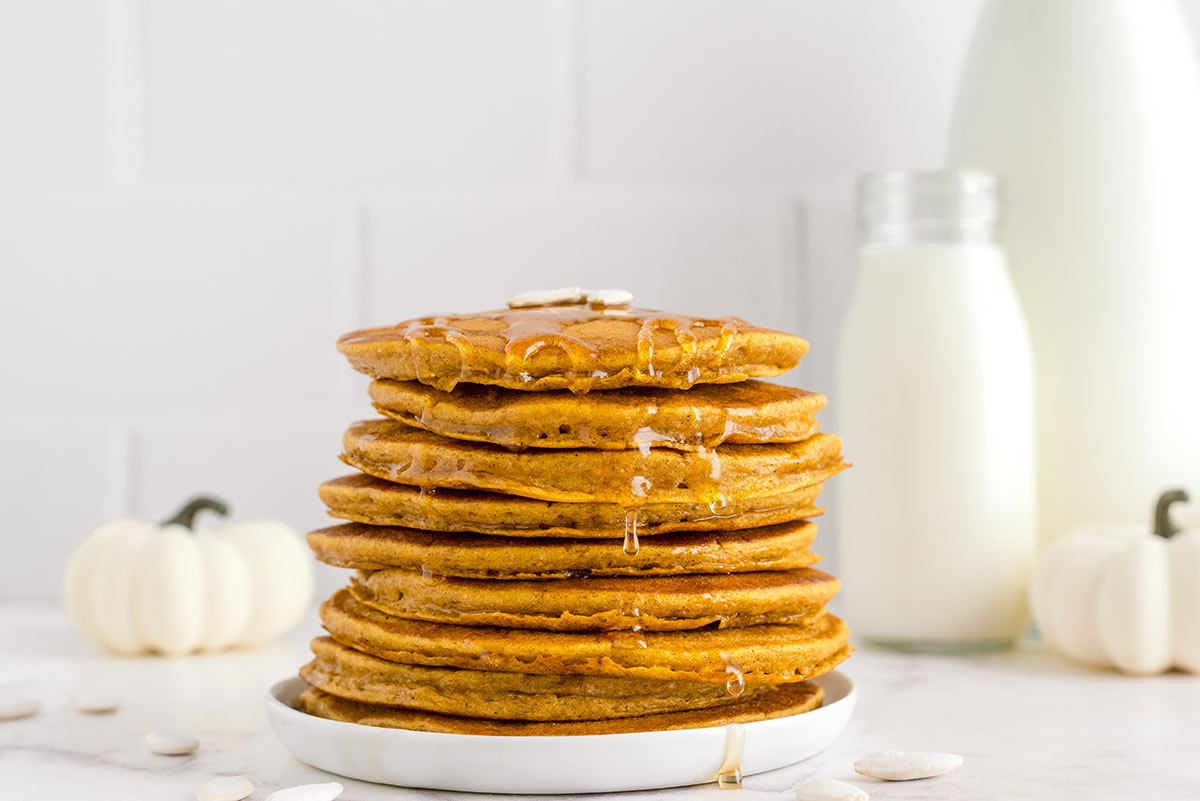 stack of fluffy pumpkin pancakes on a white plate with a white pumpkin and milk jars in the background