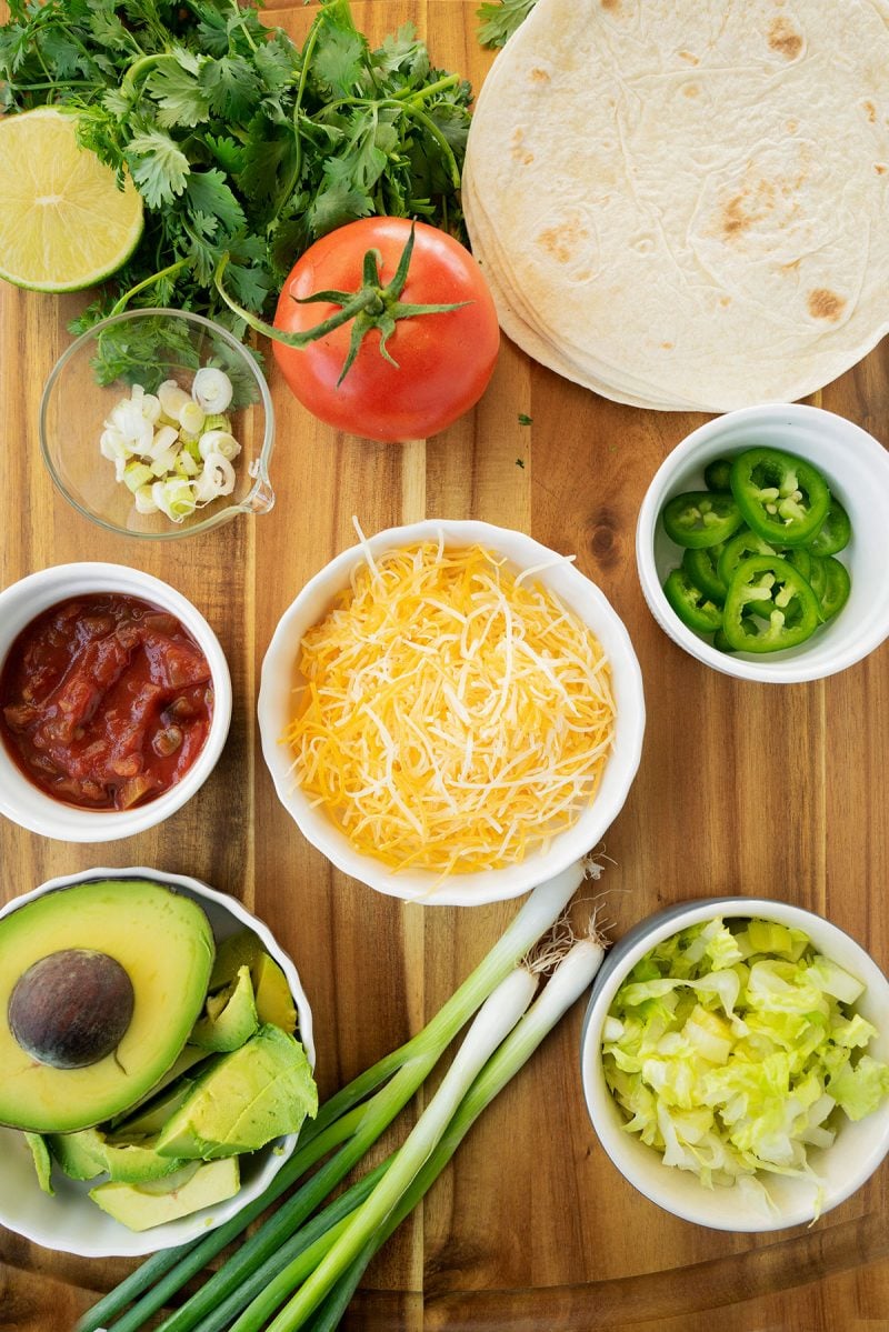 toppings on a wooden board with cheese, avocado, tomato, tortillas and scallions