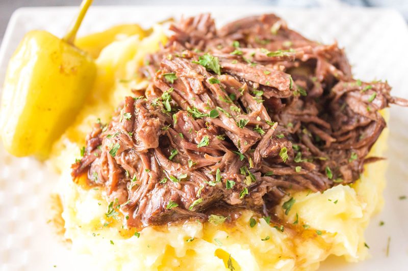 Mississippi pot roast, shredded and tender, served atop creamy mashed potatoes, garnished with chopped parsley and a whole yellow pepper on a white plate.