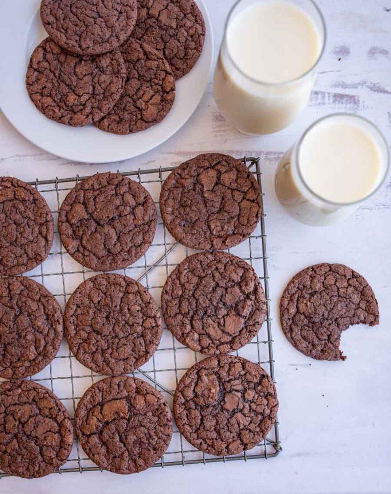 ascetic view of brownie cookies on a cooling rack with some on a plate and one cookie with a perfect bite taken out next to two glasses of milk