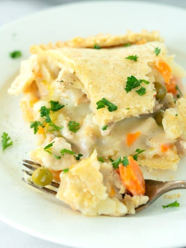 A slice of creamy chicken pot pie with carrots, peas, and a flaky crust is served on a white plate and garnished with fresh parsley. A fork rests beside the slice.