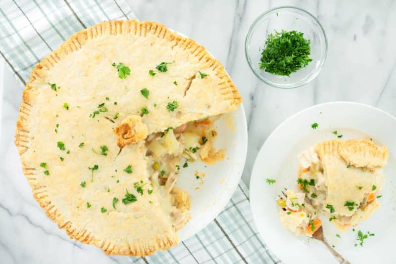 A baked pot pie with a golden crust, garnished with chopped parsley. A slice is served on a white plate nearby, showing the creamy vegetable filling. A small bowl of parsley sits above the plate on a marble surface showing what to serve with chicken pot pie.