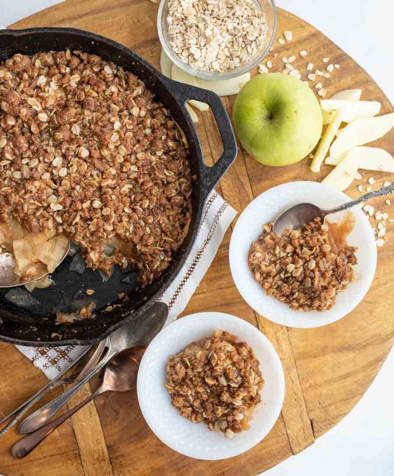two plates with apple crisp and white ice cream with the cast iron baking dish next to them with green apples on a wood block