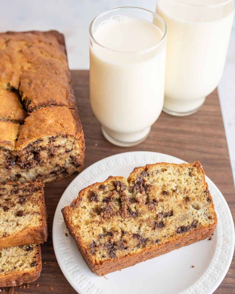 whole loaf of banana chocolate chip bread sliced and one piece on a white plate with milk to the side