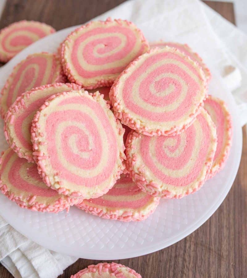 close up of a plate stacked with pink and white pinwheel cookies with a swirl design and pink sprinkles on the edges
