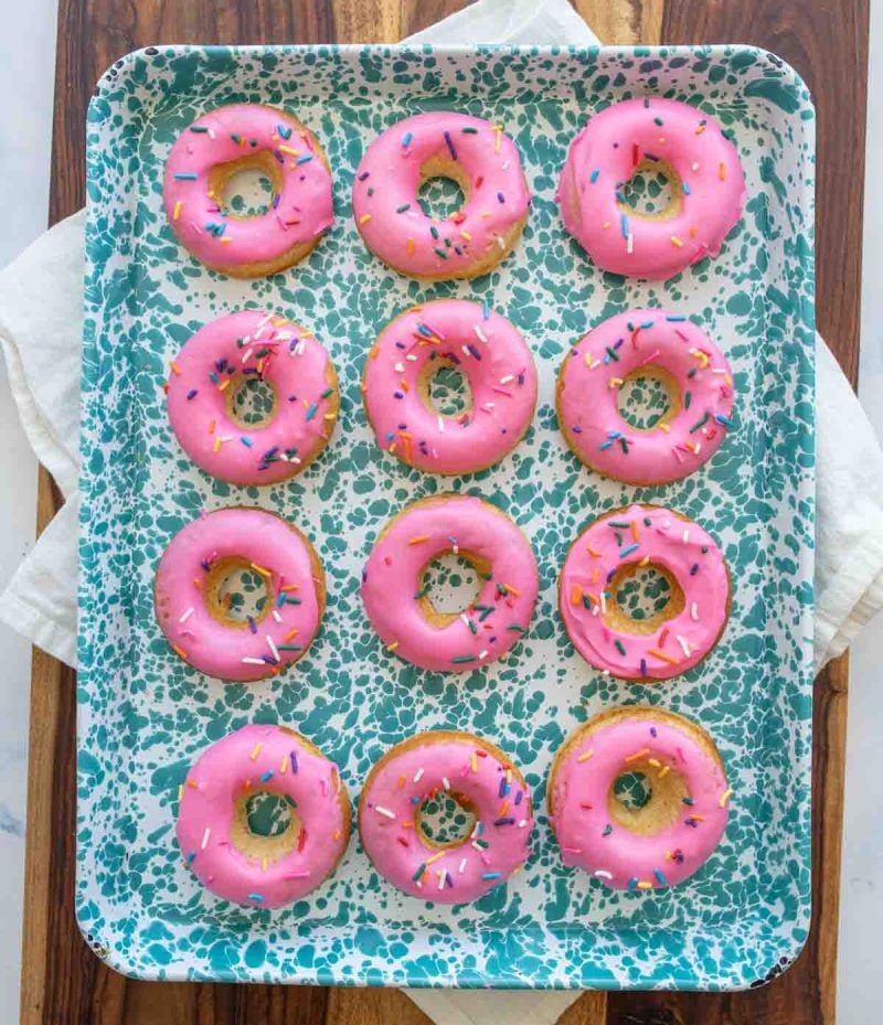 round baked donuts with pink icing and rainbow sprinkles on a baking dish