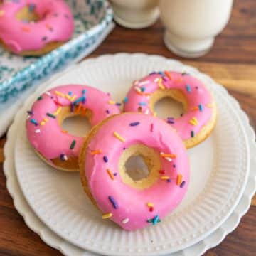 three pink iced and rainbow sprinkle round baked donuts on a plate