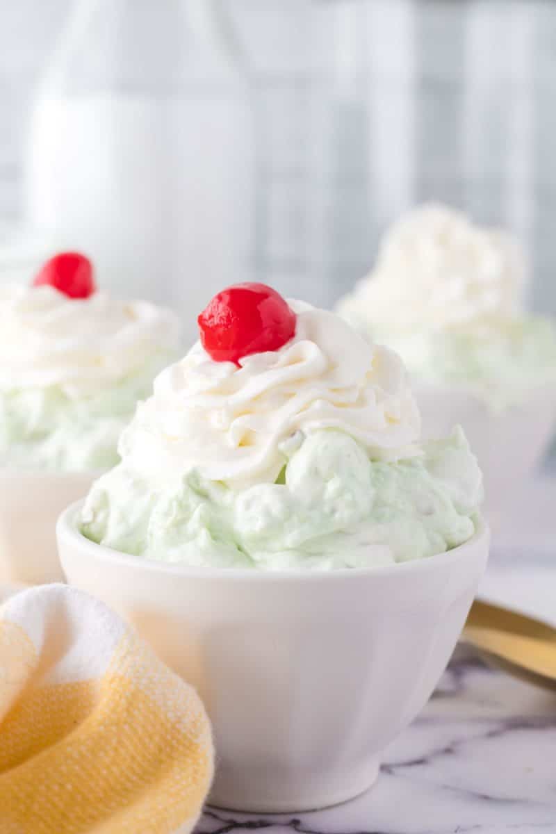 Pistachio jello salad with a cherry on top served in a white bowl.