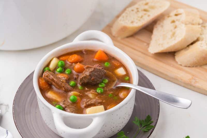 A bowl of beef stew with peas, carrots, and potatoes sits on a plate with a spoon. Slices of bread are on a wooden cutting board nearby.