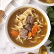 A delectable beef noodle soup recipe featuring tender pieces of beef, wide noodles, and sliced carrots in a savory broth. A wooden spoon rests nearby with fresh parsley on the side, while two golden spoons peek out at the bottom.