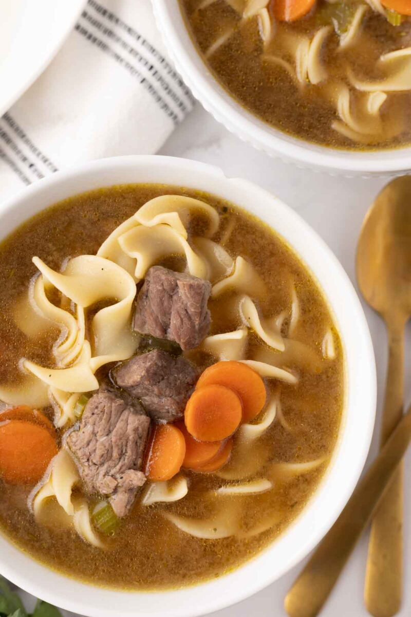 A close-up of two bowls showcasing a delicious beef noodle soup recipe, featuring tender carrots and pieces of beef, all garnished with spiral noodles. This enticing dish is served alongside a gold spoon on a white surface, partially covered by a striped cloth.