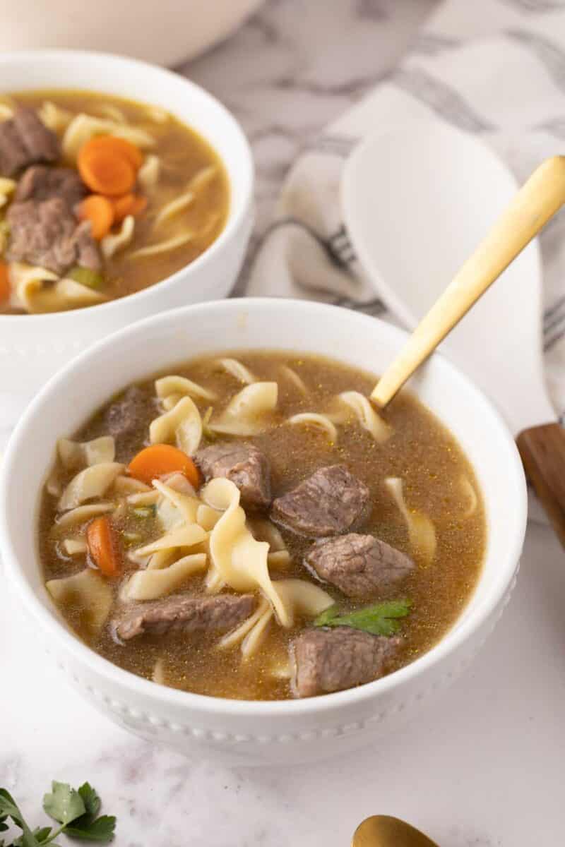 A bowl of beef noodle soup recipe with chunks of beef, sliced carrots, and wide noodles in a savory broth. A gold spoon rests in the bowl. A second bowl of soup is partially visible in the background, next to a white cloth napkin.
