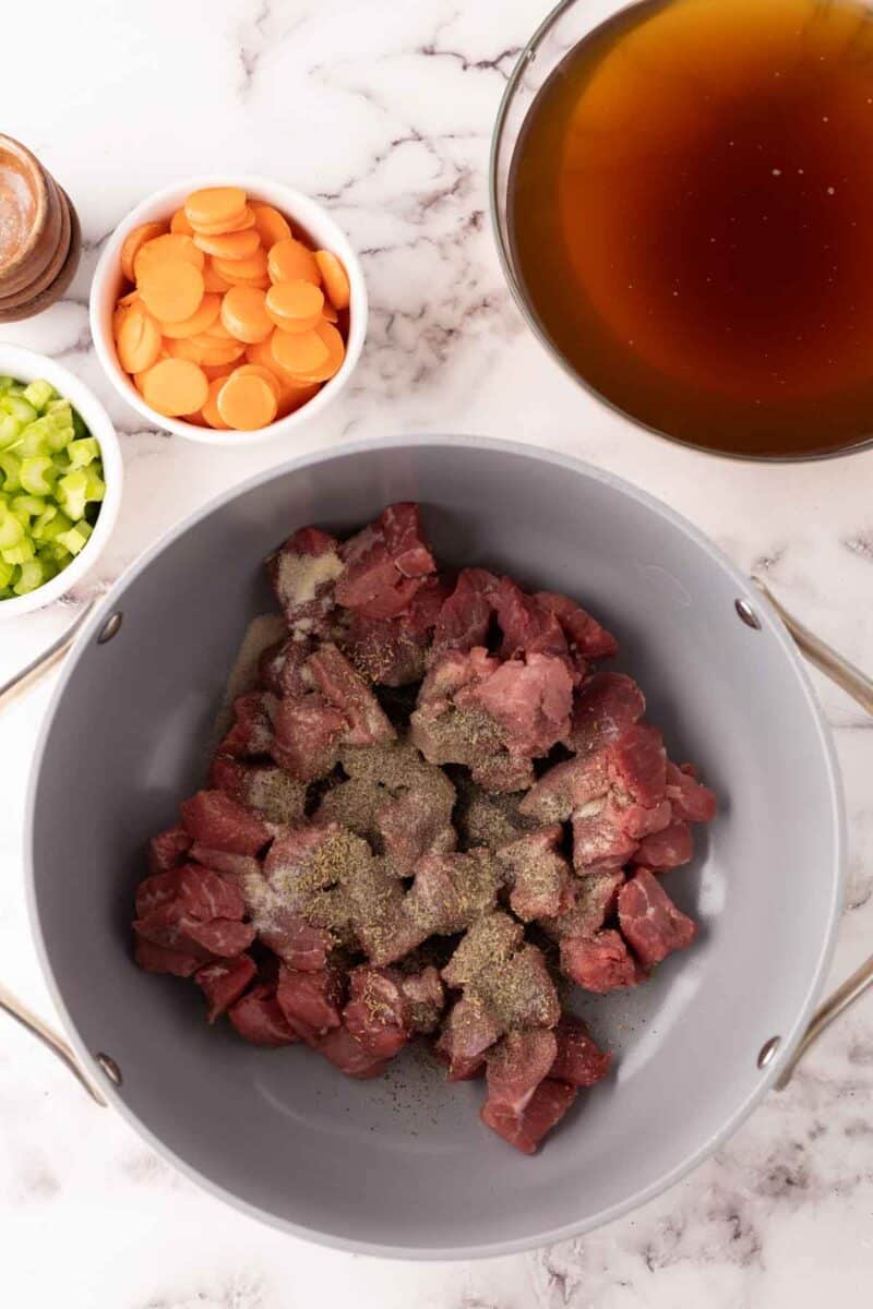 A pot filled with raw beef cubes seasoned with pepper, surrounded by bowls of sliced carrots, chopped celery, and a bowl of broth, hints at the beginnings of a hearty beef noodle soup recipe. The scene is set on a marble countertop.