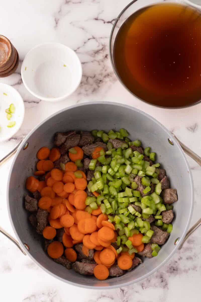 A pot on the marble countertop holds chopped carrots, celery, and pieces of meat essential for a beef noodle soup recipe. Nearby, two small white bowls—one with spices and the other empty—accompany a larger bowl filled with dark liquid.