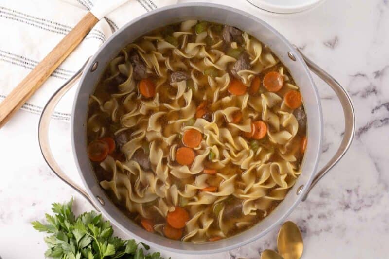 A pot of savory beef noodle soup with wide egg noodles, sliced carrots, and chunks of beef graces the marble countertop. A wooden spoon, a striped napkin, fresh parsley, and gold-colored utensils accompany this delightful beef noodle soup recipe.