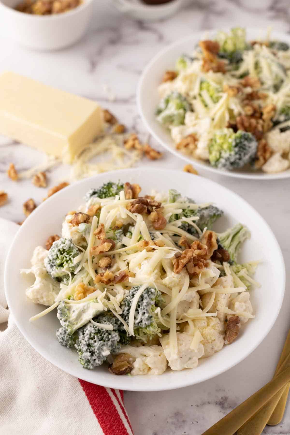 A plate of creamy broccoli cauliflower salad topped with shredded cheese and walnuts. The salad is served on a white dish with a block of cheese in the background. A fork and red-striped napkin are placed beside the plate.