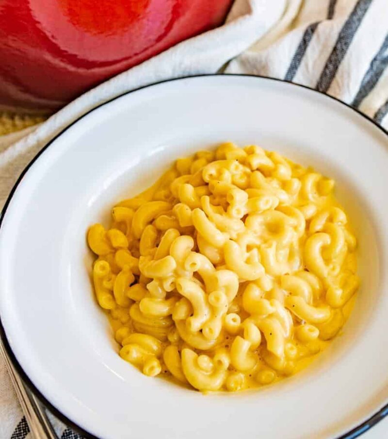 A white bowl filled with creamy kids mac and cheese sits on a striped cloth next to a red pot.