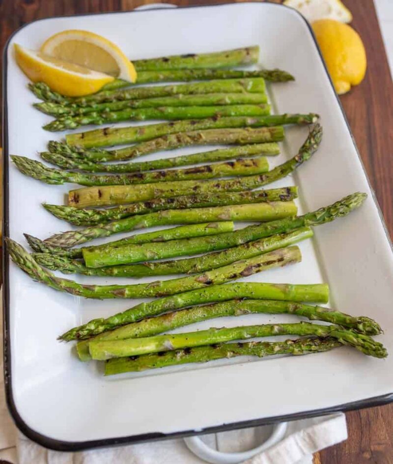 Grilled asparagus spears arranged in a white rectangular dish, garnished with two lemon wedges in the corner. The dish sits on a wooden surface.
