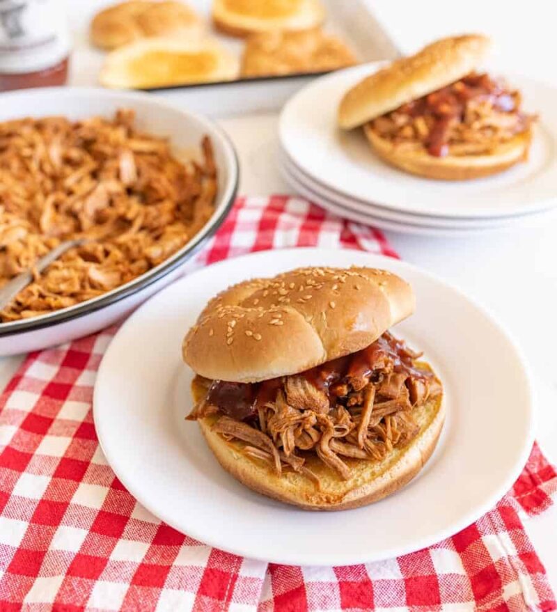 A pulled chicken sandwich with barbecue sauce on a sesame bun sits on a white plate. Nearby are more sandwiches, a bowl of shredded chicken, and instant pot pulled pork tenderloin on a baking sheet of buns, all on a red-and-white checkered cloth.