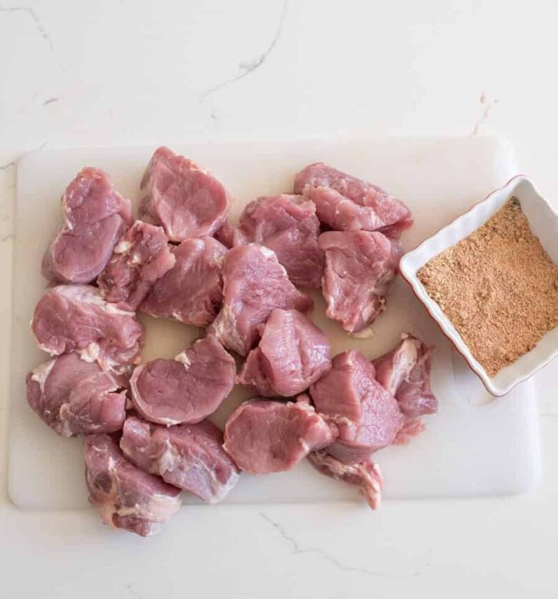 Chunks of raw pork, perfect for instant pot pulled pork tenderloin, are set on a white cutting board next to a small square dish filled with brown seasoning mix, all atop a white countertop.