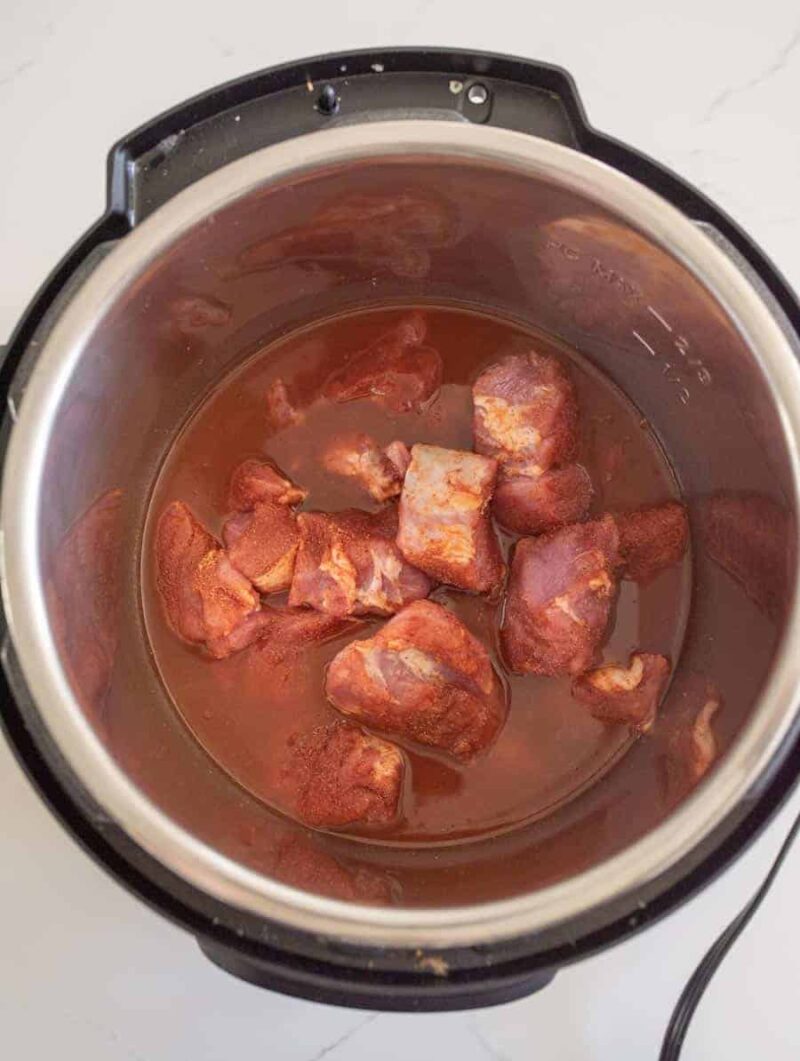 Chunks of raw, seasoned meat partially submerged in liquid inside the metal pot of an Instant Pot, ready to be cooked for tender pulled pork tenderloin, viewed from above.